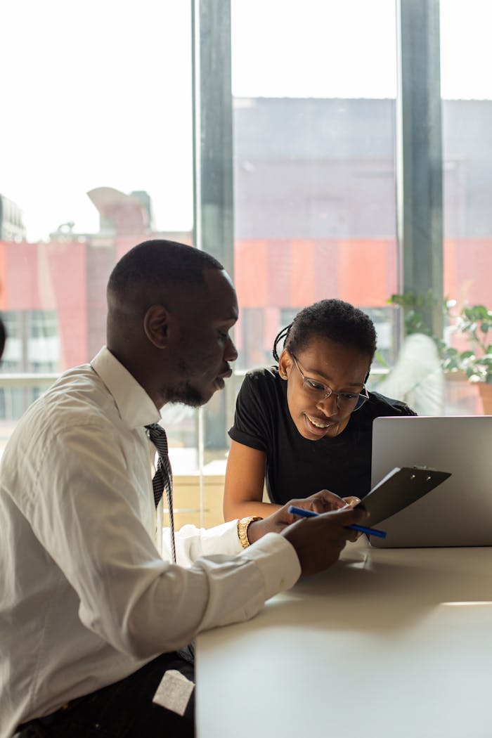Two professionals collaborate over a laptop and clipboard in a modern office setting.